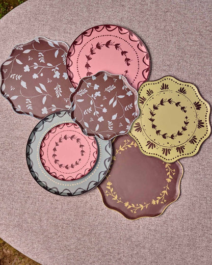 Decorative plates some with scalloped edges are arranged in a cluster on a round table. The plates feature various patterns and colors including pink, brown, yellow, and gray, with some showing floral motifs and others displaying geometric or lace-like borders.