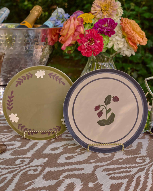 A styled outdoor table scene with decorative plates, a hammered metal ice bucket with bottles, a glass, flowers, and a bowl of cherries. The central focus appears to be the decorative ceramic plates, with one plate standing upright and another leaning behind it on a tablecloth.