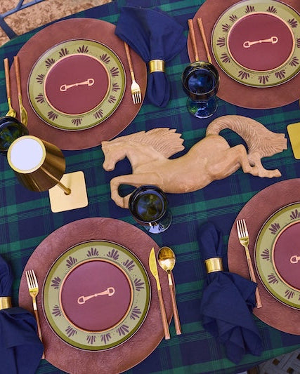 An overhead view of a rustic outdoor table set for four, featuring a green and blue plaid tablecloth, matching plates with gold-rimmed edges, gold cutlery, dark blue napkins, and a central wooden horse figurine.