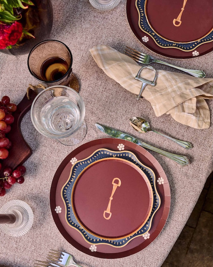 An overhead styled table setting featuring a round tableware set with deep reddish-brown plates, blue and gold wavy borders, silver flatware, beige napkins, and grapes on a linen tablecloth.