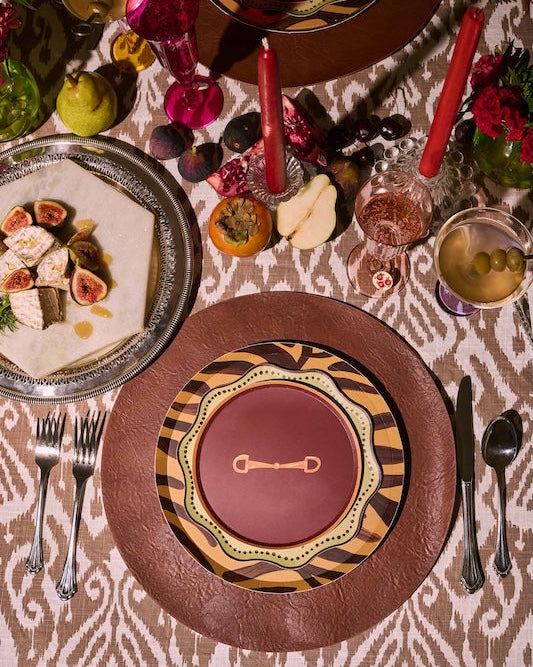 An overhead styled table setting featuring a round brown leather charger plate layered with a brown and black patterned plate, topped with a deep red center plate. The table is decorated with red taper candles, glasses, fresh figs, sliced apples, and a silver platter holding fig and white bean wraps on a patterned tablecloth.