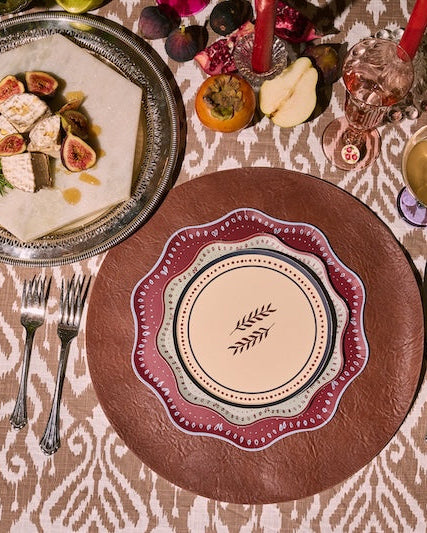 An overhead styled table setting featuring a central place setting with a brown textured charger plate, a cream and red patterned dinner plate, silver cutlery, and a serving platter of figs and fish, all arranged on a beige and brown patterned tablecloth.