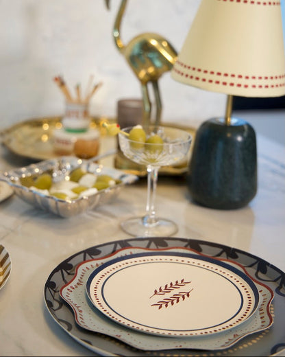 A styled tabletop setting featuring a decorative dinner plate with a red leaf motif in the center.  cream-colored table lamp with red dotted trim and a dark green base stands next to a gold flamingo figurine and a glass coupe filled with olives.