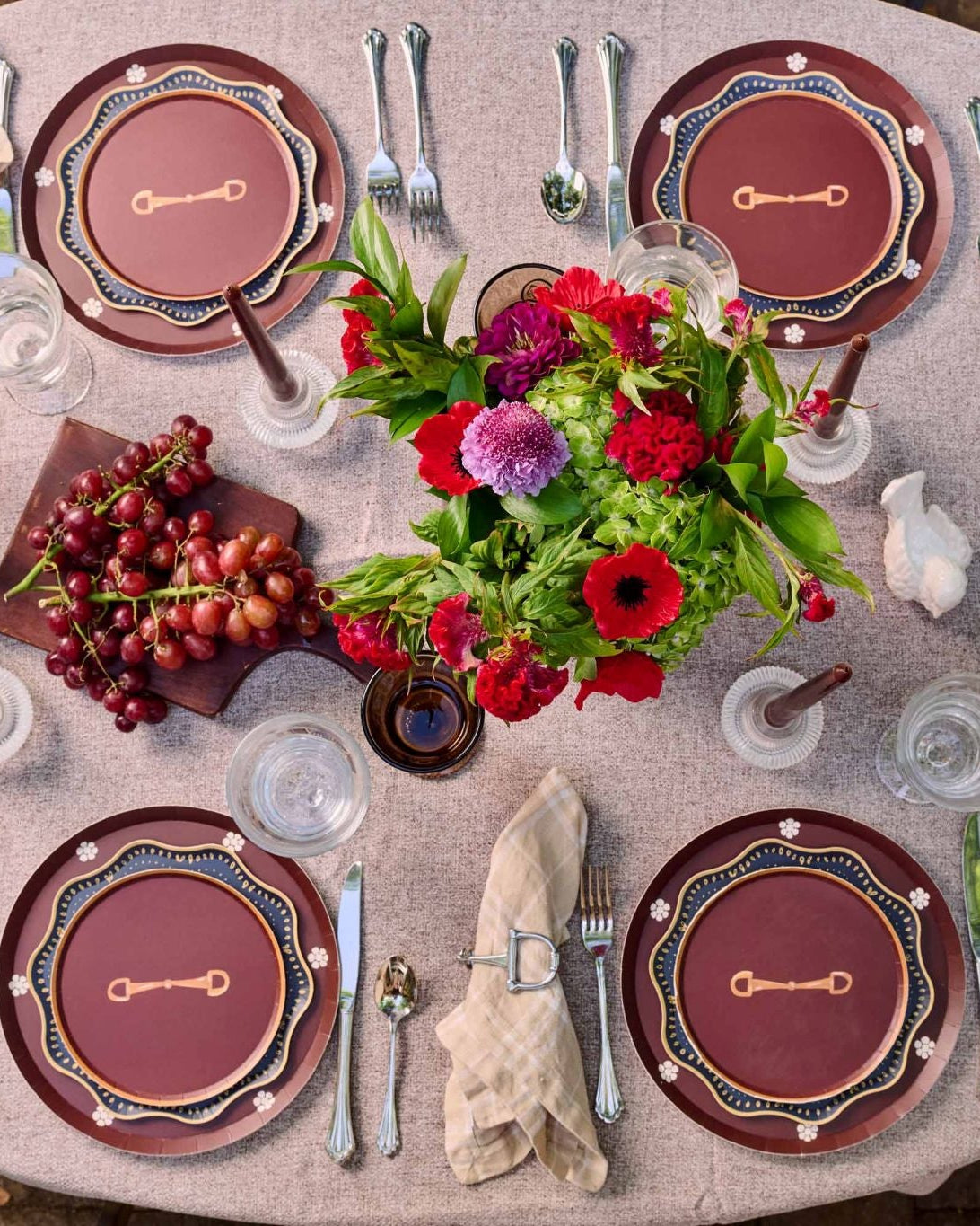 An overhead view of a beautifully set round dining table with 4 place settings, silver cutlery, folded cream napkins, clear drinking glasses, a central floral centerpiece, and a small wooden board of red grapes on a textured beige tablecloth.