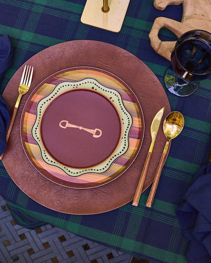 A styled outdoor table setting featuring rustic brown plates with scalloped multicolor borders, navy blue napkins in gold rings, gold cutlery, and a green and blue plaid tablecloth.