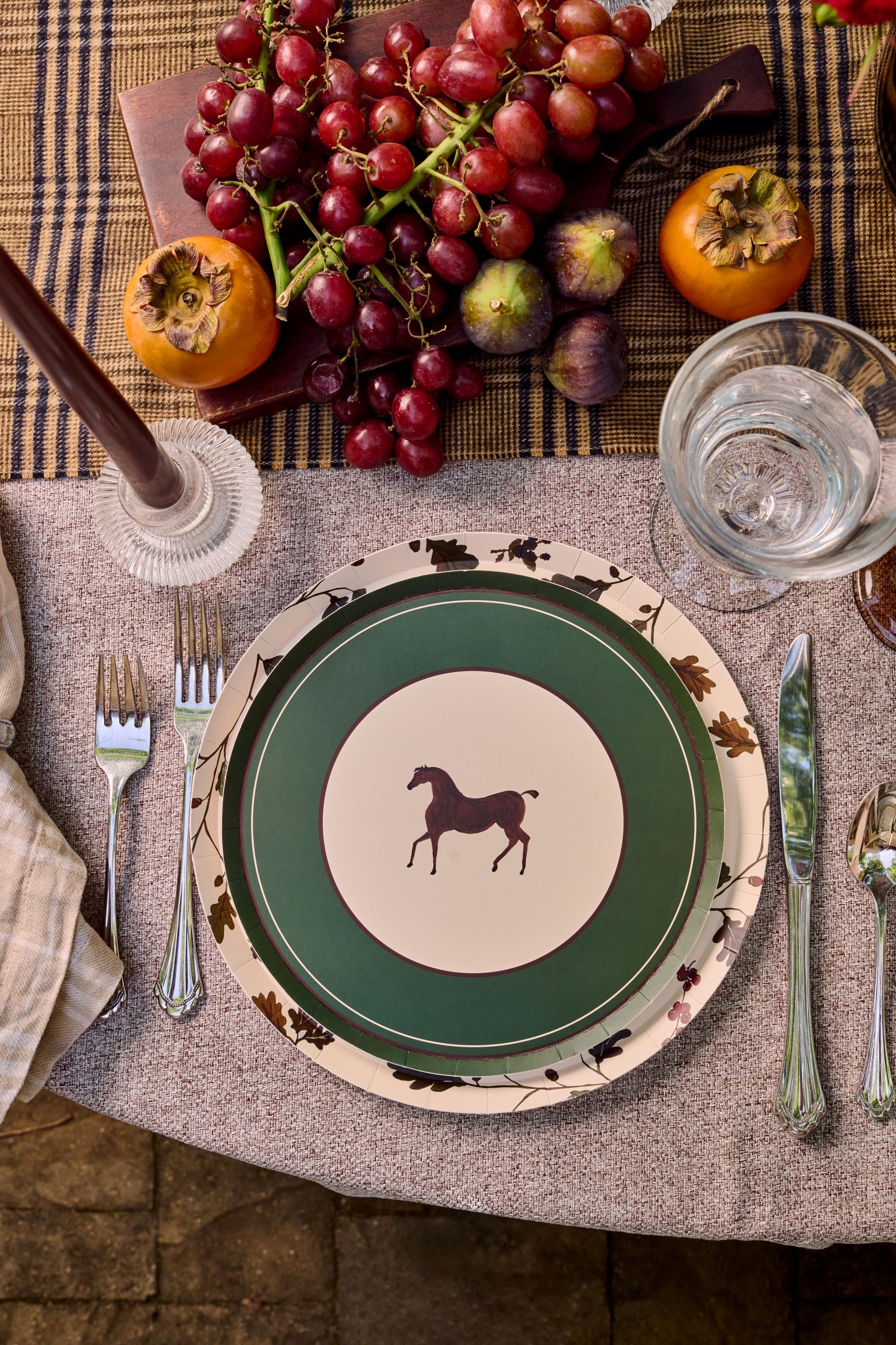 A styled tabletop setting featuring a green and cream ceramic plate with a horse silhouette, layered with a floral-patterned charger plate, alongside silver cutlery, a glass of water, and autumn fruits on a plaid runner.