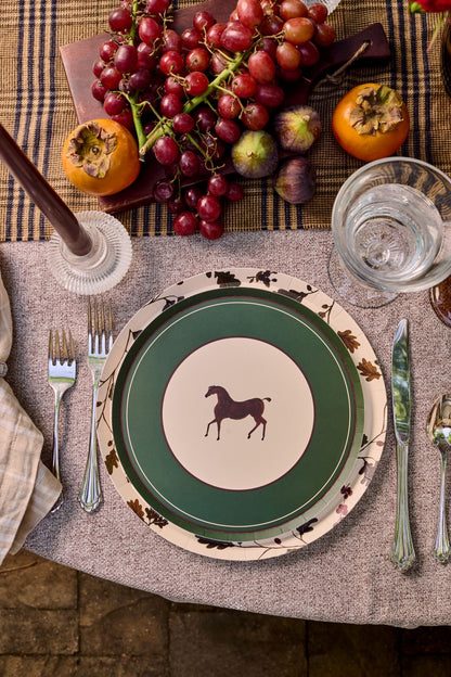 A styled tabletop setting featuring a green and cream ceramic plate with a horse silhouette, layered with a floral-patterned charger plate, alongside silver cutlery, a glass of water, and autumn fruits on a plaid runner.