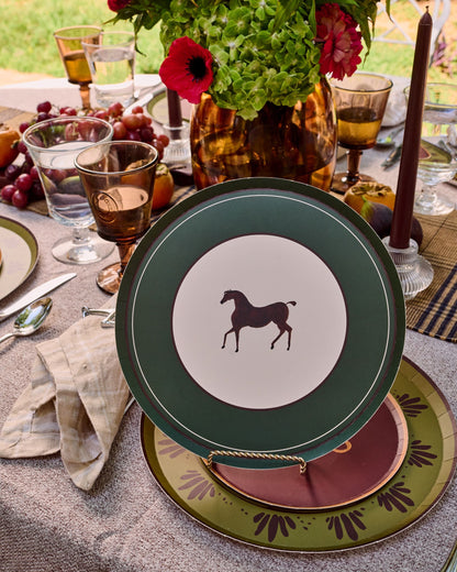 Two decorative serving plates displayed on a table outdoors, with a floral arrangement, ice bucket, and bottles in the background. One plate is sage green with a cream center and a dark horse silhouette, and the other is dusty red with a gold equestrian bit motif.