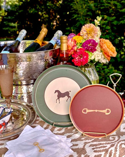 Two decorative serving plates displayed on a table outdoors, with a floral arrangement, ice bucket, and bottles in the background. One plate is sage green with a cream center and a dark horse silhouette, and the other is dusty red with a gold equestrian bit motif.