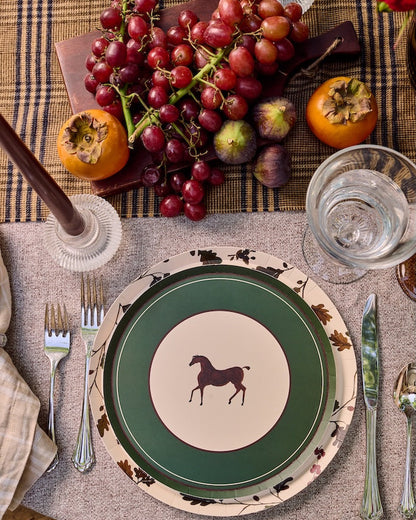 A styled tabletop setting featuring a green and cream ceramic plate with a horse silhouette, layered with a floral-patterned charger plate, alongside silver cutlery, a glass of water, and autumn fruits on a plaid runner.